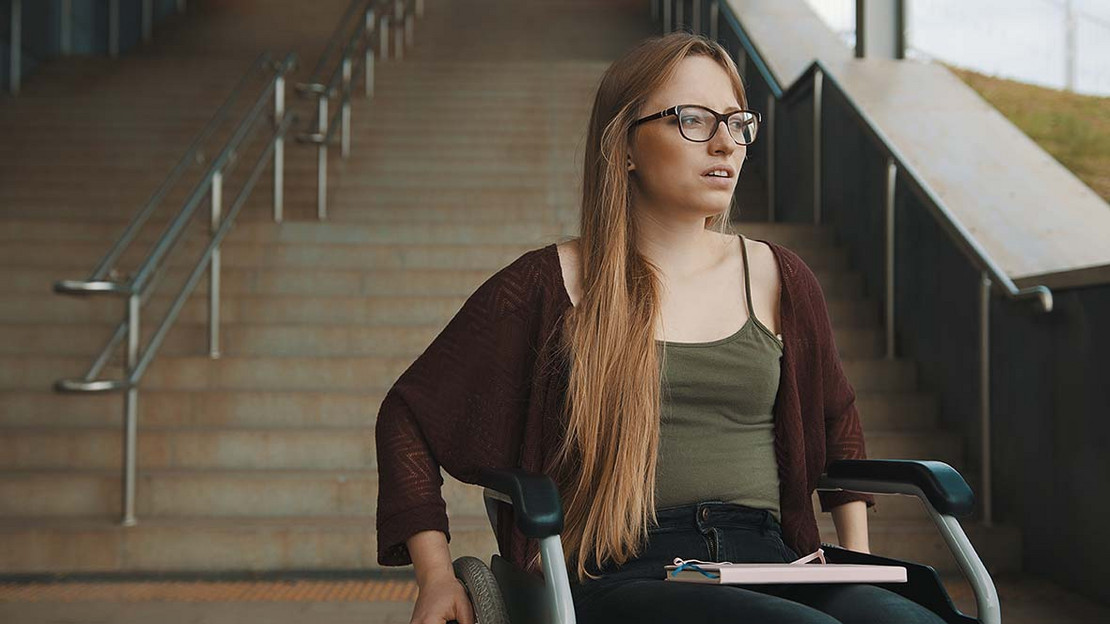 Junge Frau in einem Rollstuhl steht am Ende einer Treppe. 