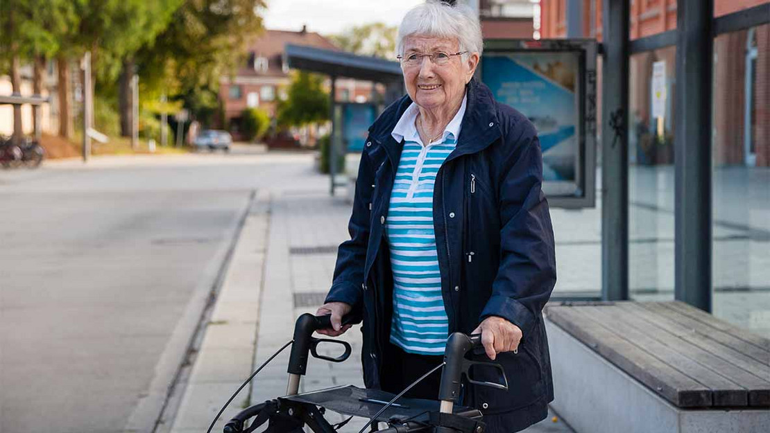 Ältere Frau mit einem Rollator steht an einer Bushaltestelle. 