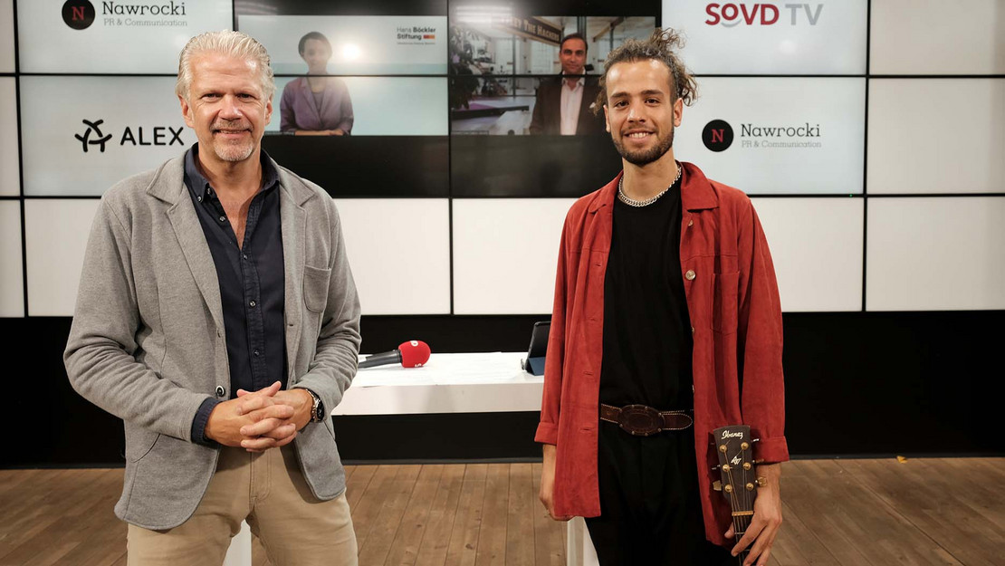 Zwei Männer im Studio. Im Hintergrund eine Bildschirmwand mit zwei Gesichtern. 