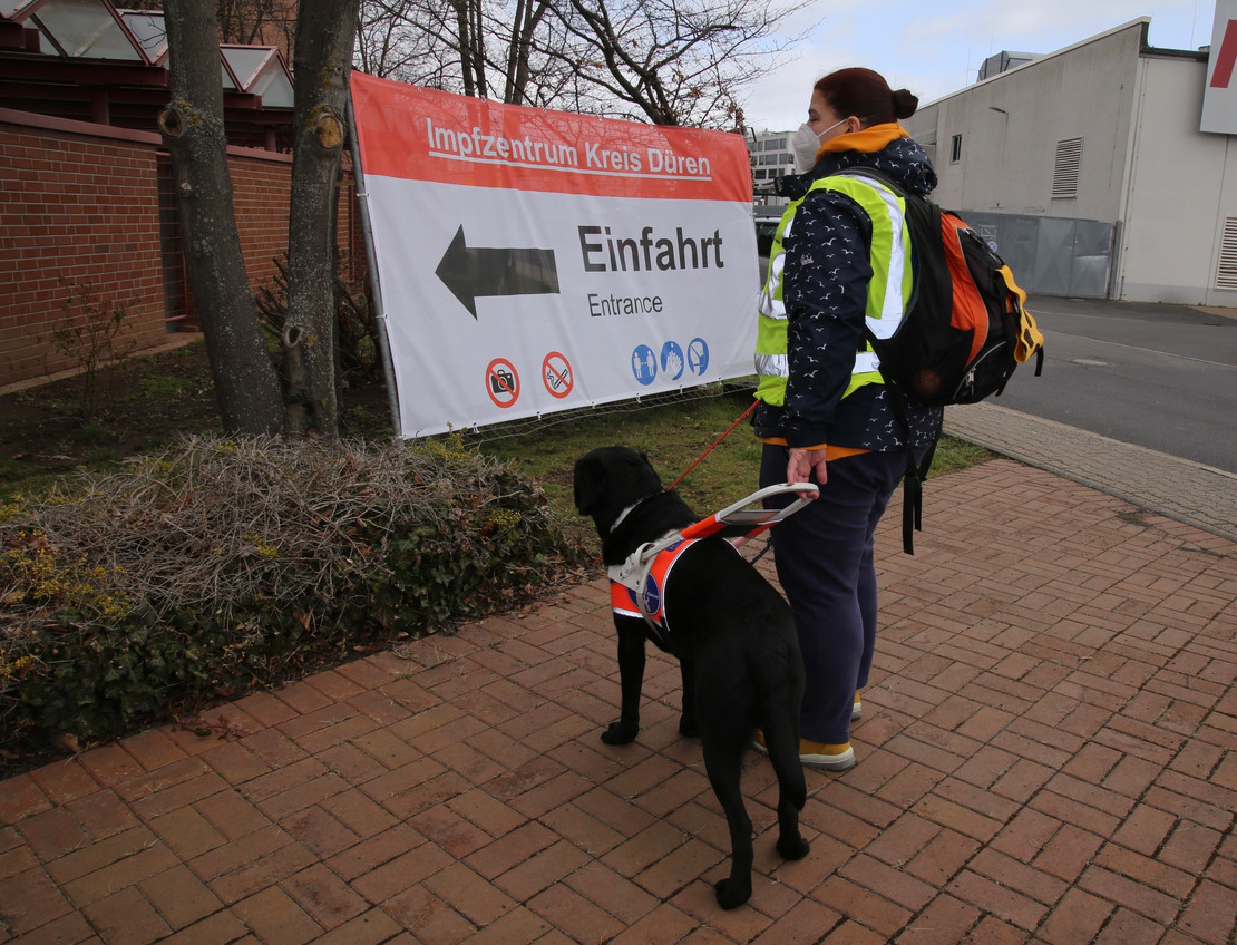 Frau mit Assistenzhund vor einem Impfzentrum. 