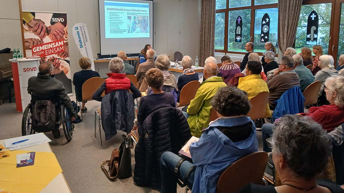 Volle Plätze in einem Veranstaltungssaal. Ein Referent spricht vor einer Leinwand. 